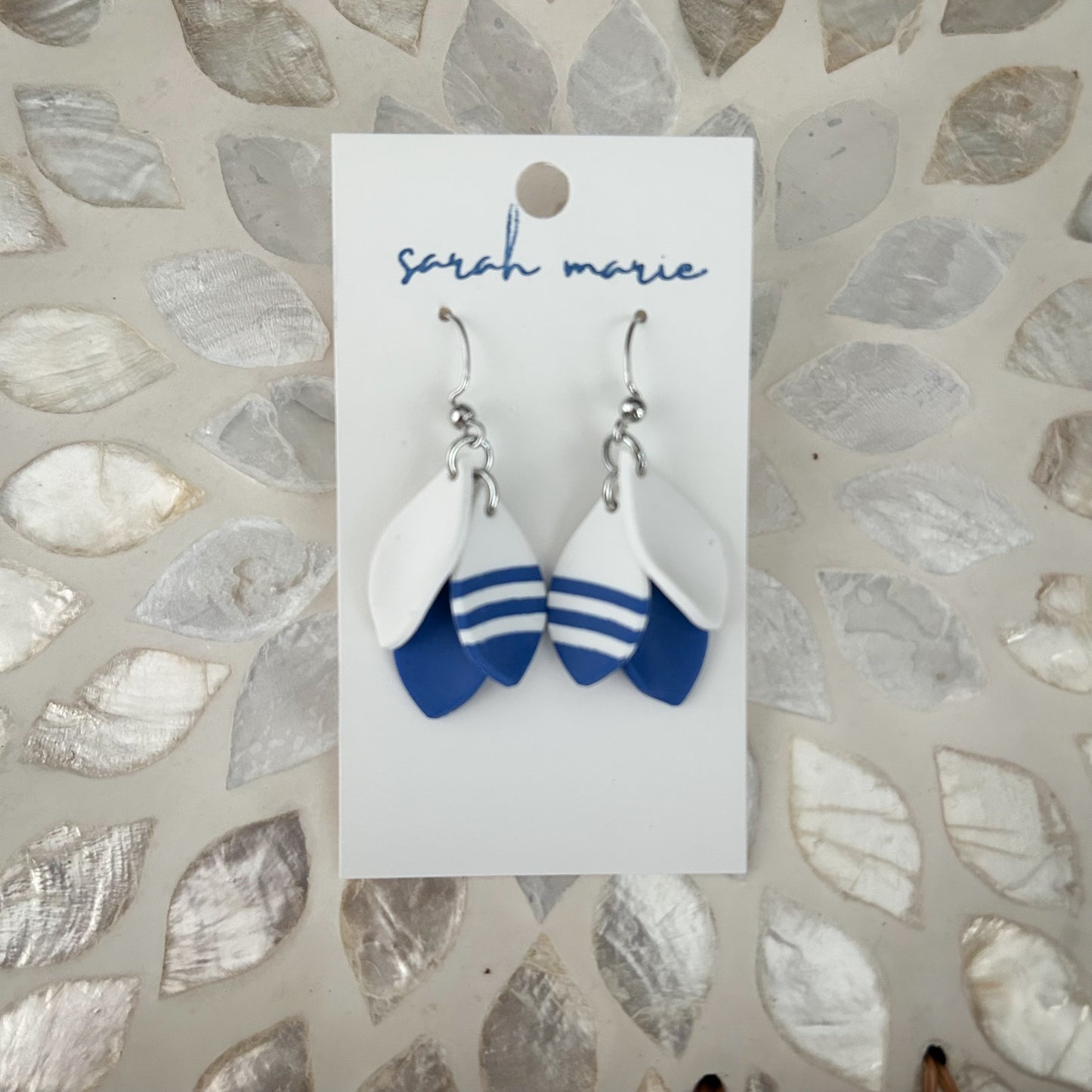 Pair of blue and white 1.5” dangle hook earrings on a 'sarah marie' branded card against a textured stone background