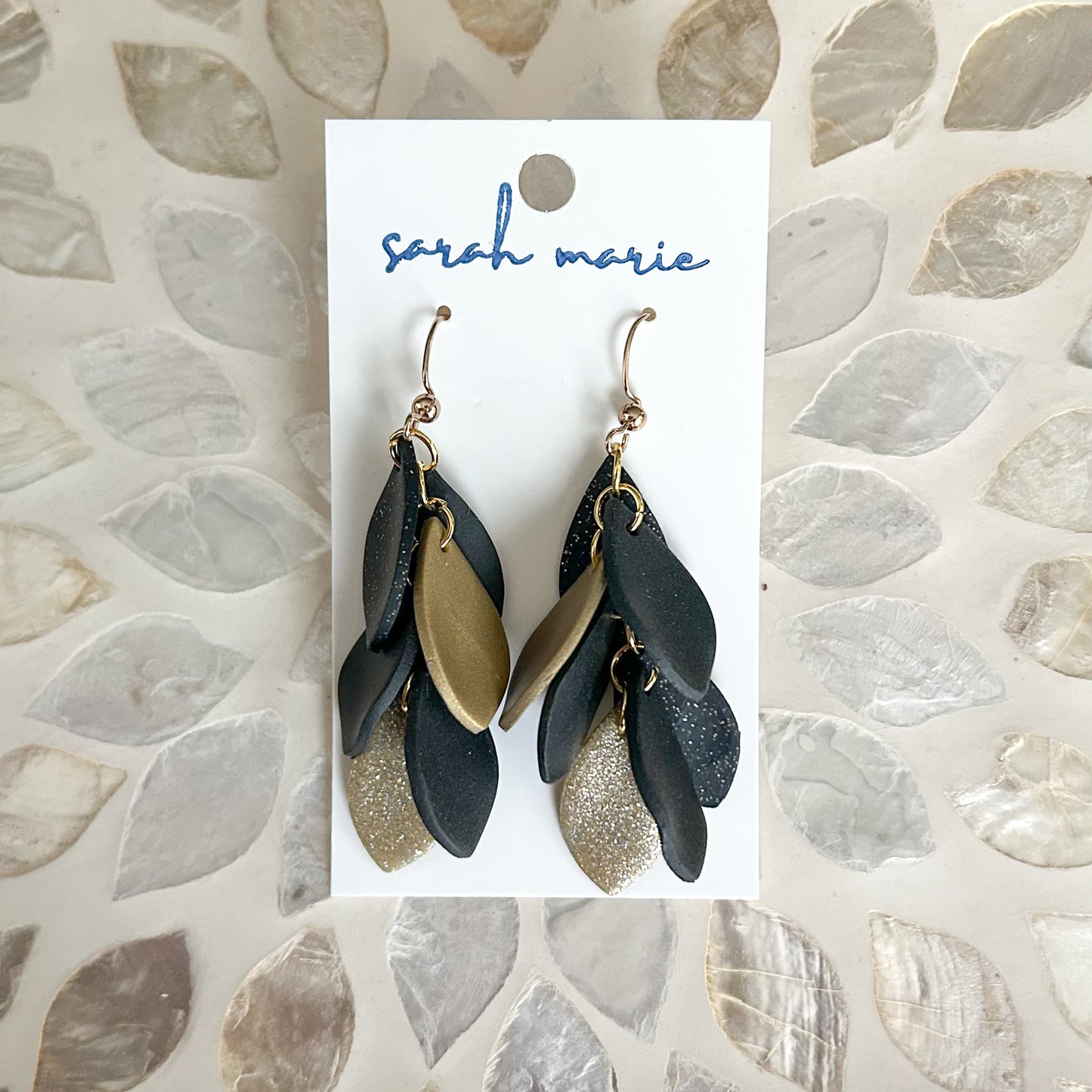 Black and gold dangle leaf-shaped earrings on a 'Sarah Marie' branded card against a textured stone background.