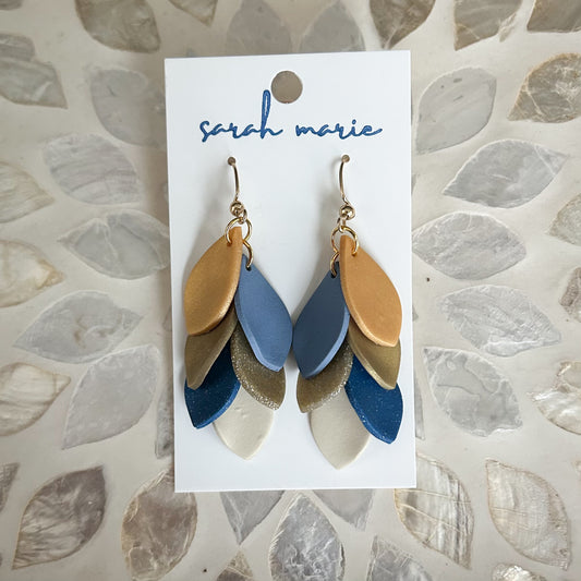 Pair of gold and blue leaf-shaped earrings on a 'Sarah Marie' branded card against a textured stone background.