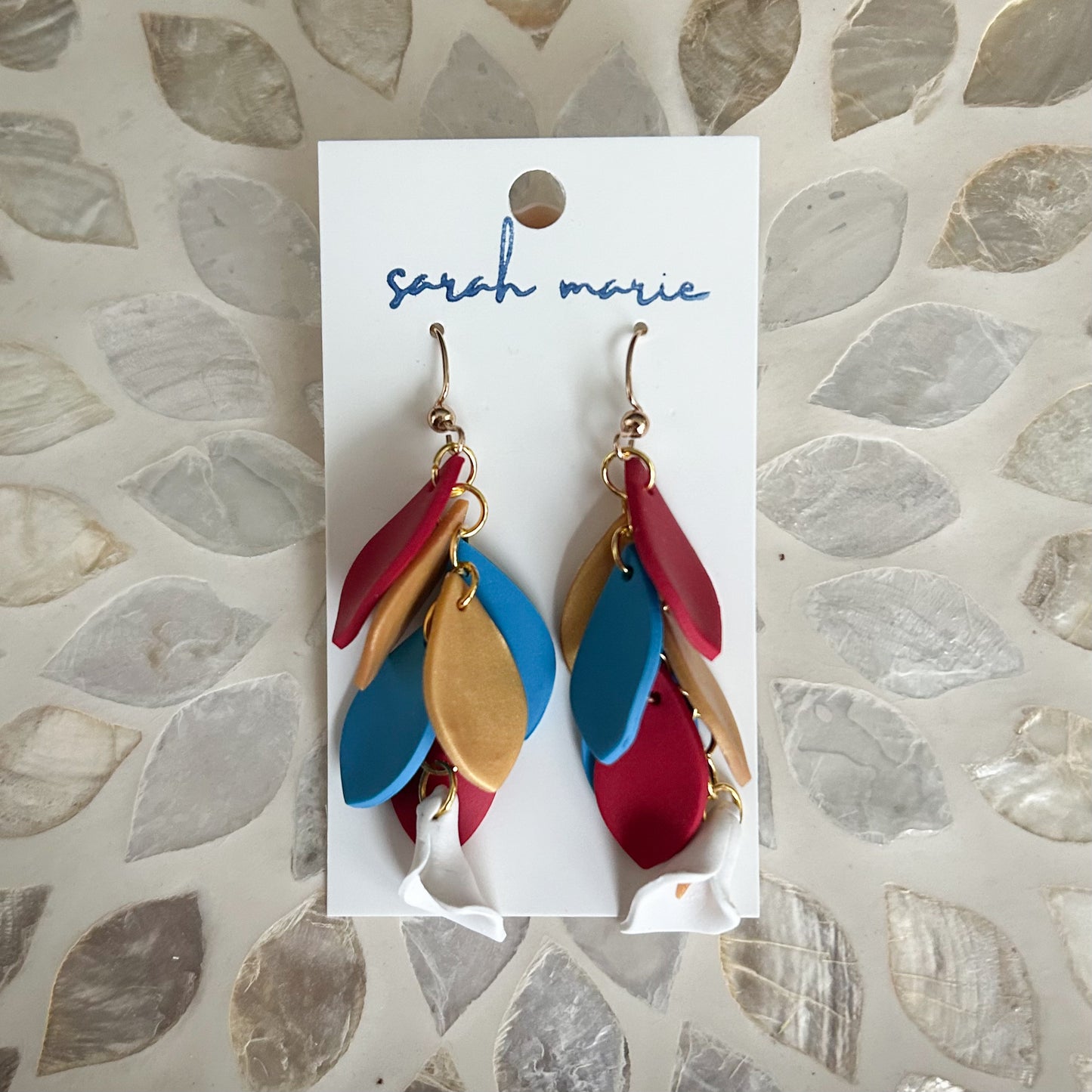 Colorful red blue and gold feather-shaped hook dangle earrings on a 'sarah marie' branded card against a textured background.