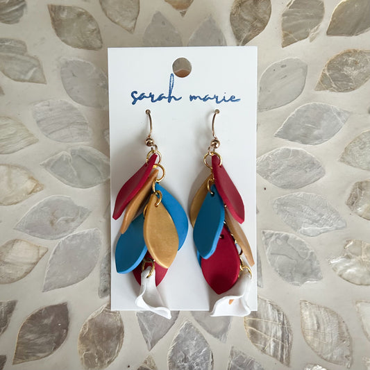 Colorful red blue and gold feather-shaped hook dangle earrings on a 'sarah marie' branded card against a textured background.