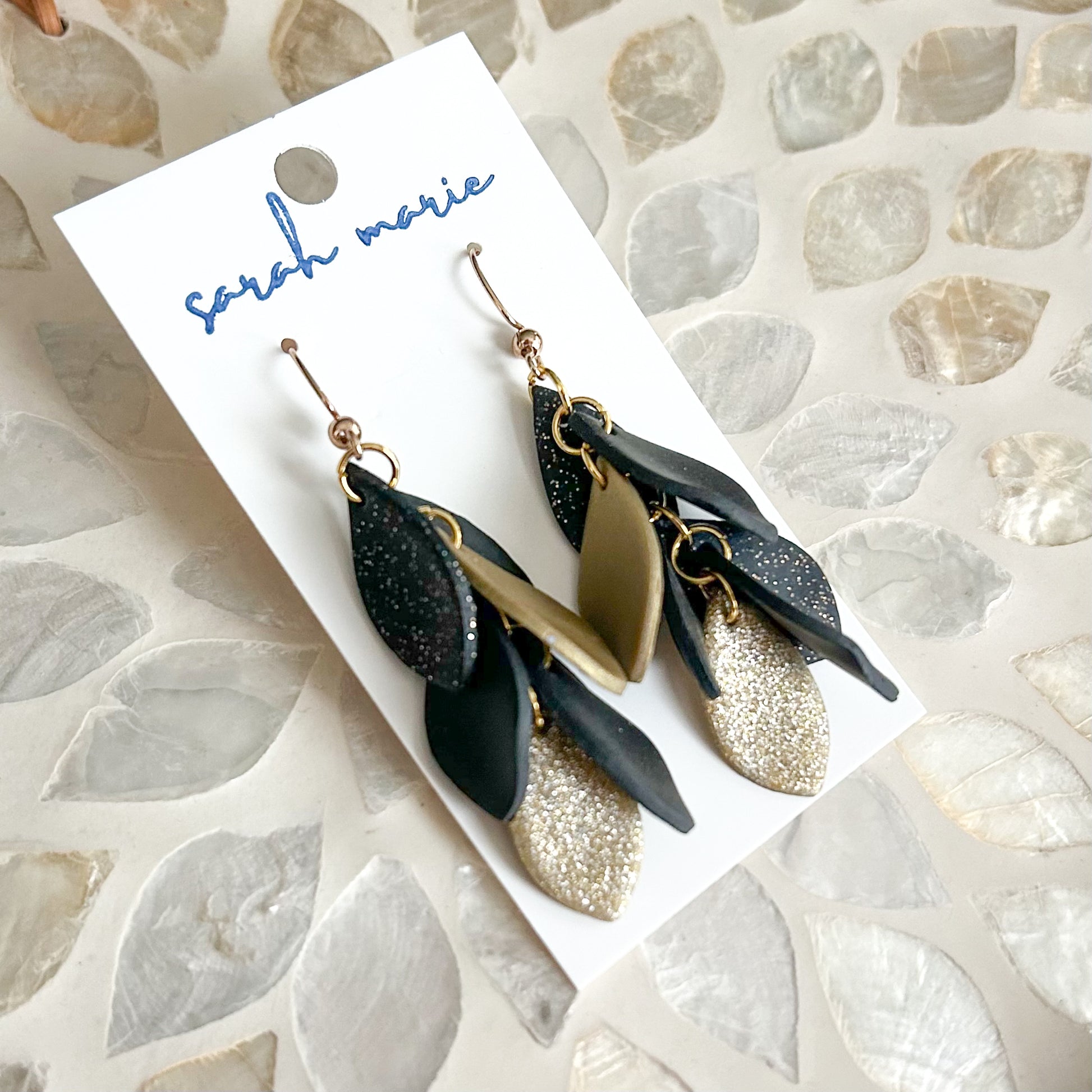 Black and gold leaf shaped earrings on a 'sarah marie' branded card against a textured stone background