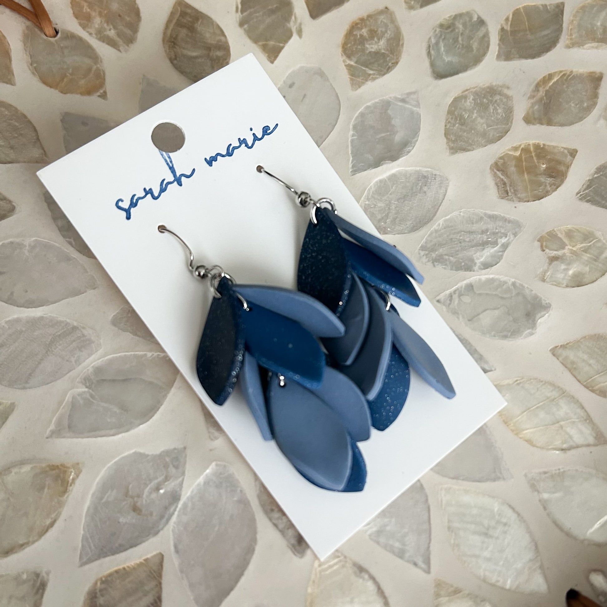Blue leaf-shaped earrings on a 'Sara Marie' branded card against a stone-patterned background
