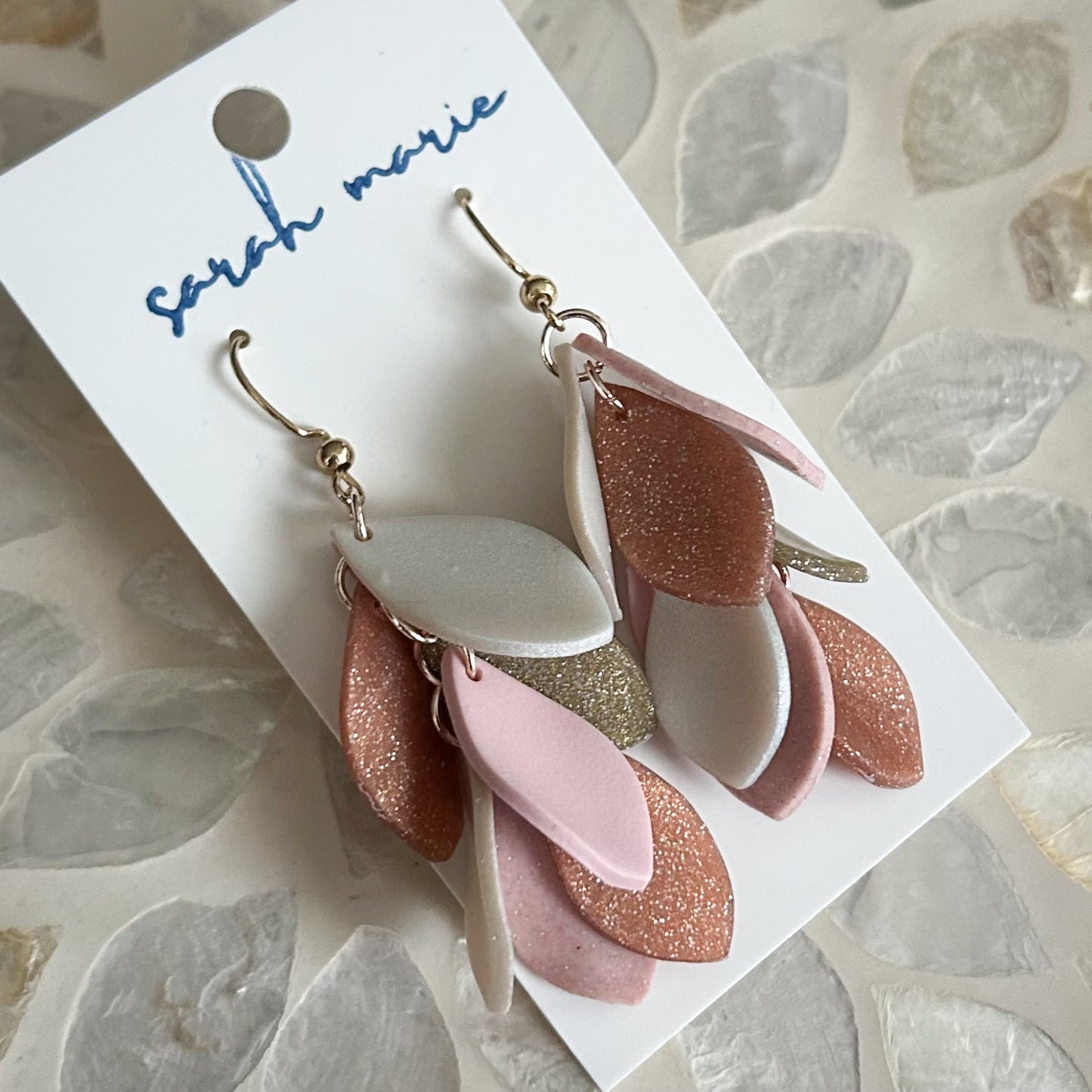 Pair of pink, rose gold and gold dangle hook earrings on a 'Sarah Marie' branded card against a stone surface.
