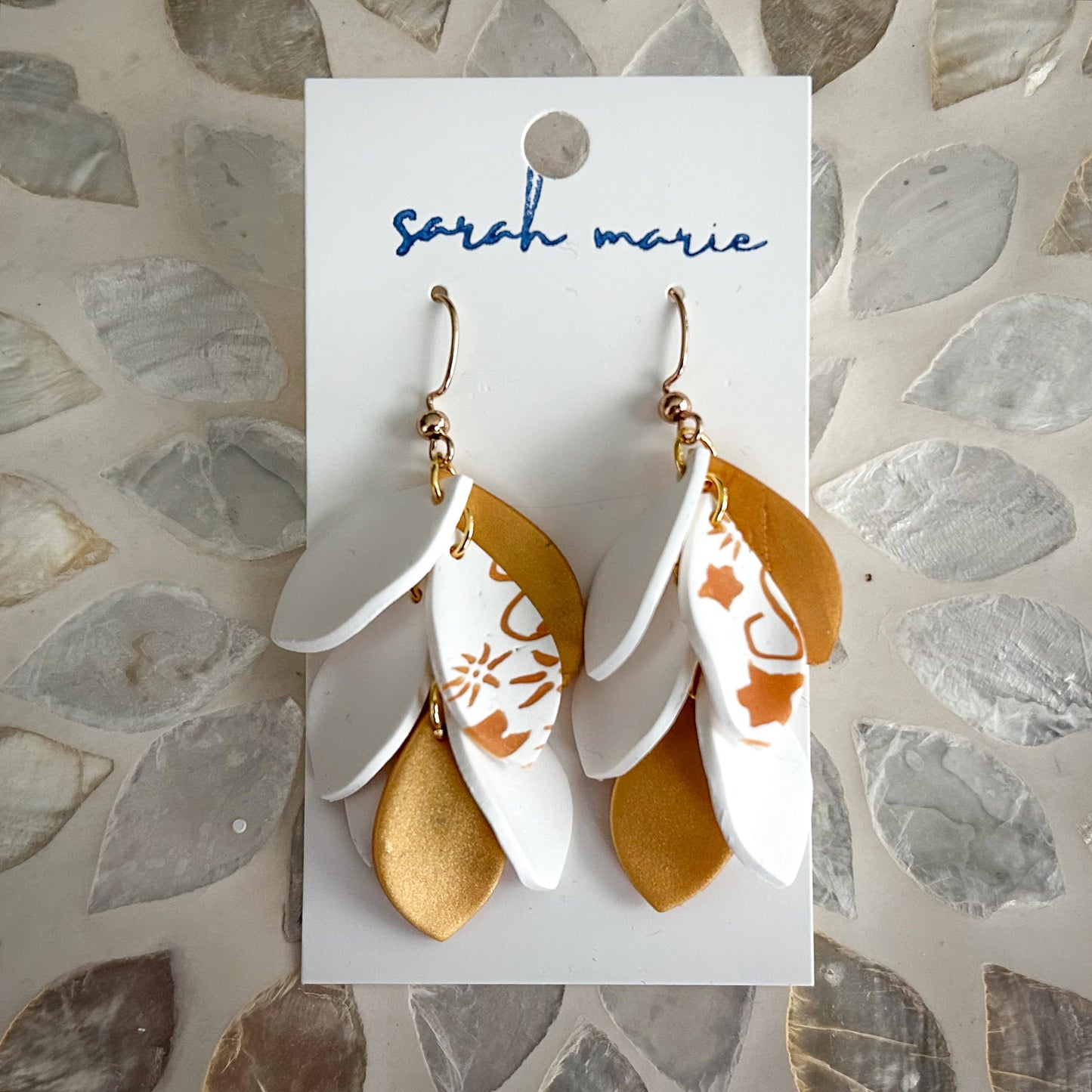 Pair of gold and white leaf-shaped dangle hook earrings on on a 'sarah marie' branded card against a stone background.