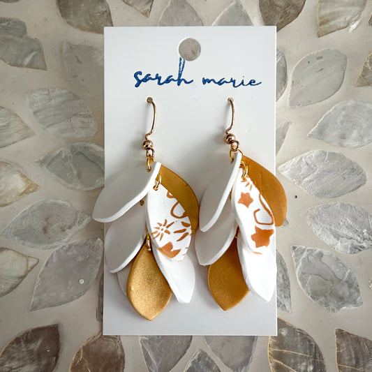 Pair of gold and white leaf-shaped dangle hook earrings on on a 'sarah marie' branded card against a stone background.