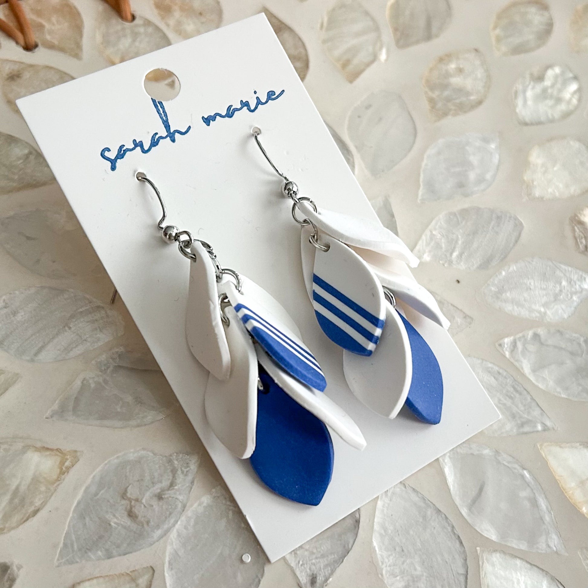 Pair of blue and white dangle hook earrings on a 'Sarah Marie' branded card against a textured stone background.