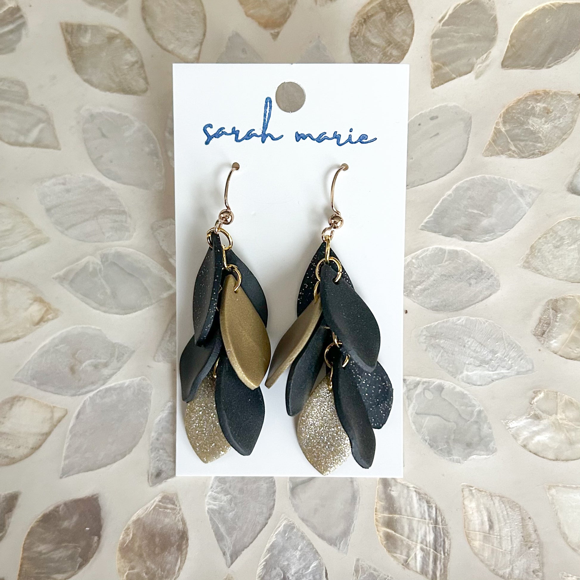 Black and gold dangle leaf-shaped earrings on a 'Sarah Marie' branded card against a textured stone background.