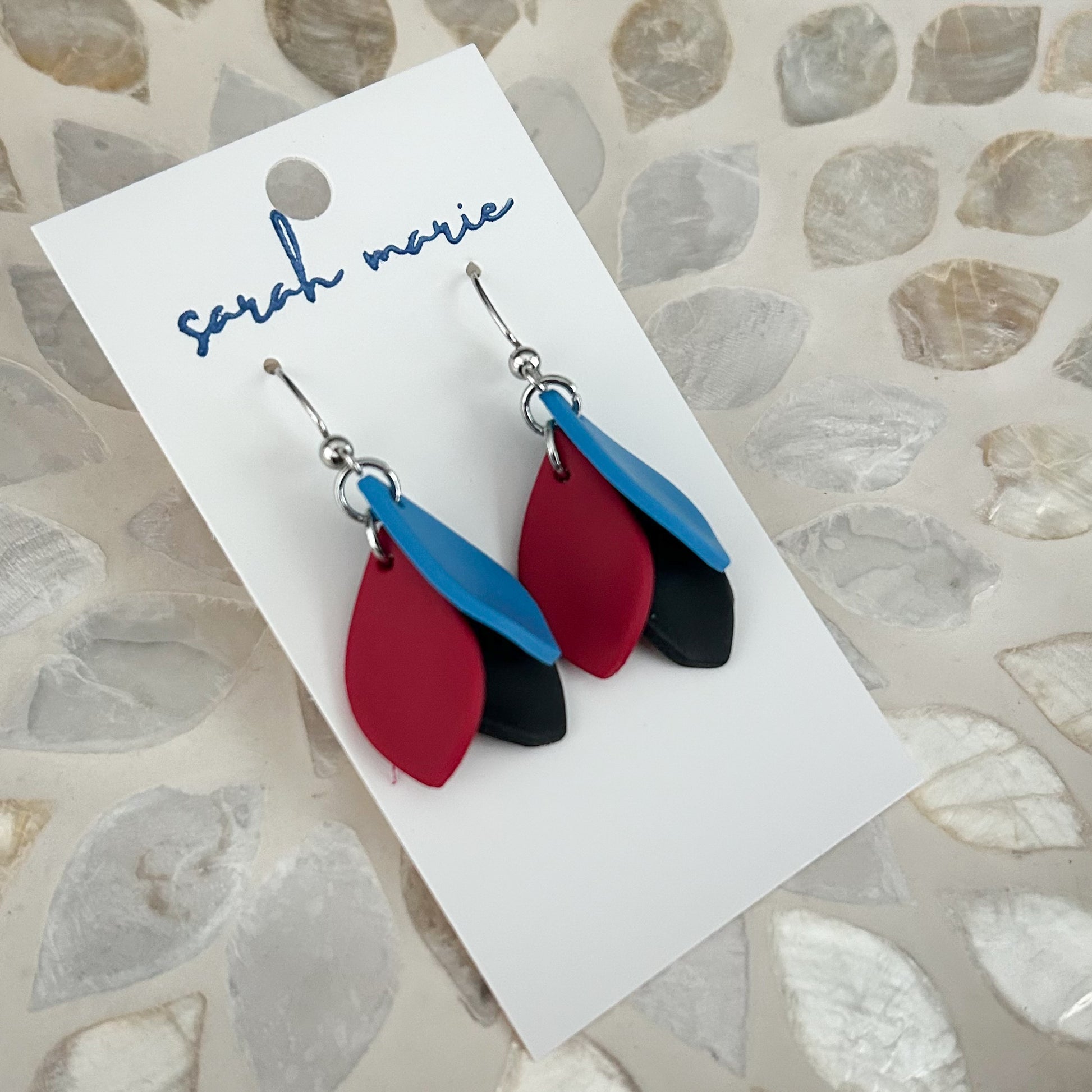 Red blue and black leaf-shaped earrings on a 'sarah marie' branded card against a stone-patterned background.