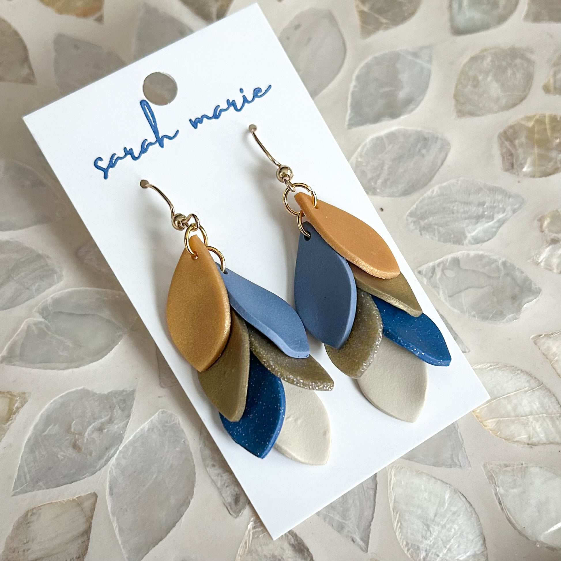 Blue and gold leaf-shaped dangle hook earrings on a 'sarah marie' branded card against a stone-patterned background